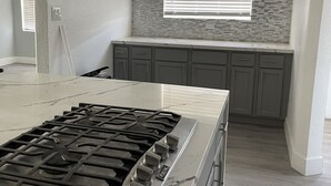 A modern kitchen with a central island, grey cabinets, a stone backsplash, and a window with blinds. - Cozy ranch house with indoor pool (Norco)