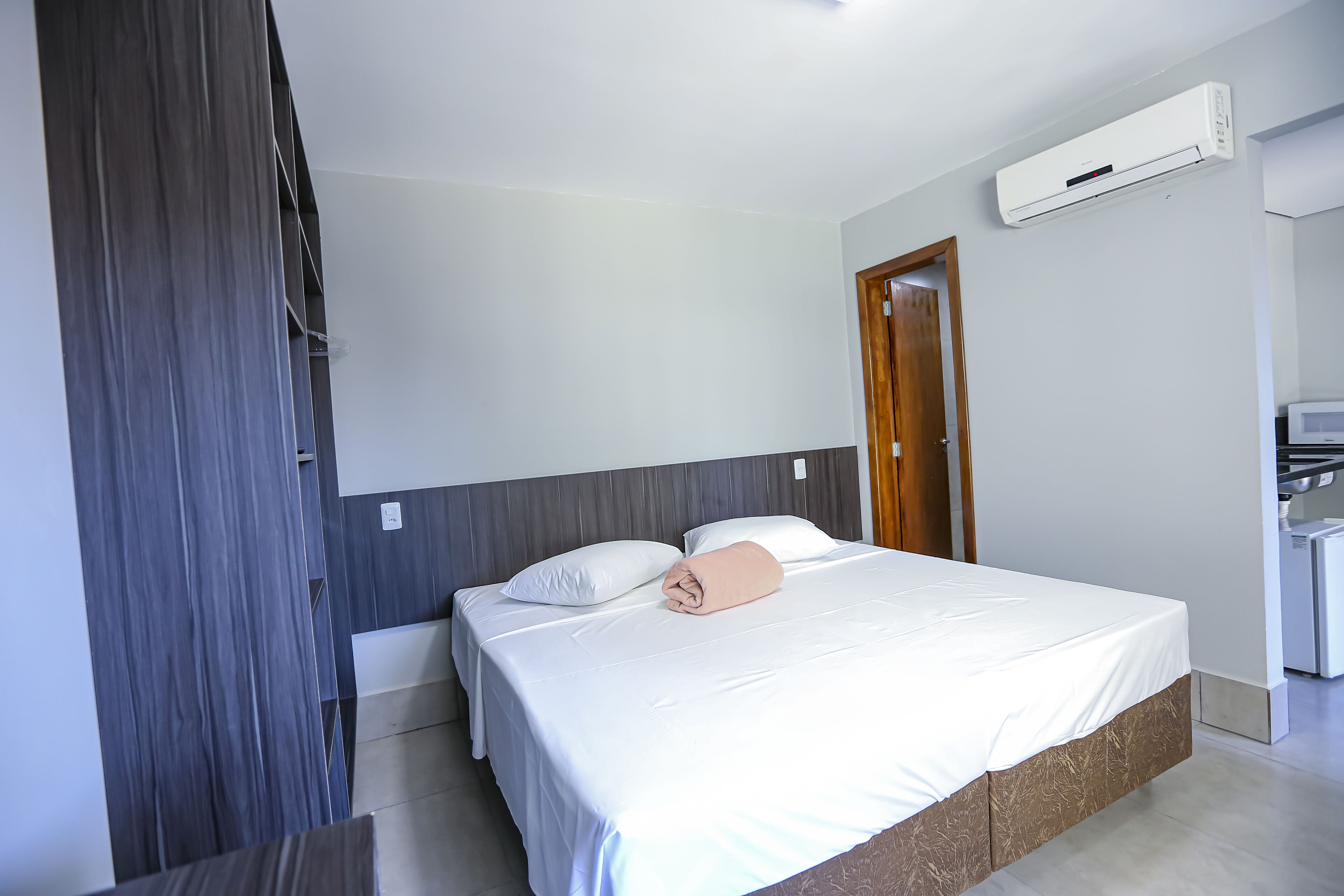 A hotel room with a bed, a wooden door, a white air conditioning unit, and a small kitchenette area.