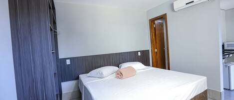 Quarto de hotel com cama, porta de madeira, ar condicionado branco e uma pequena cozinha.