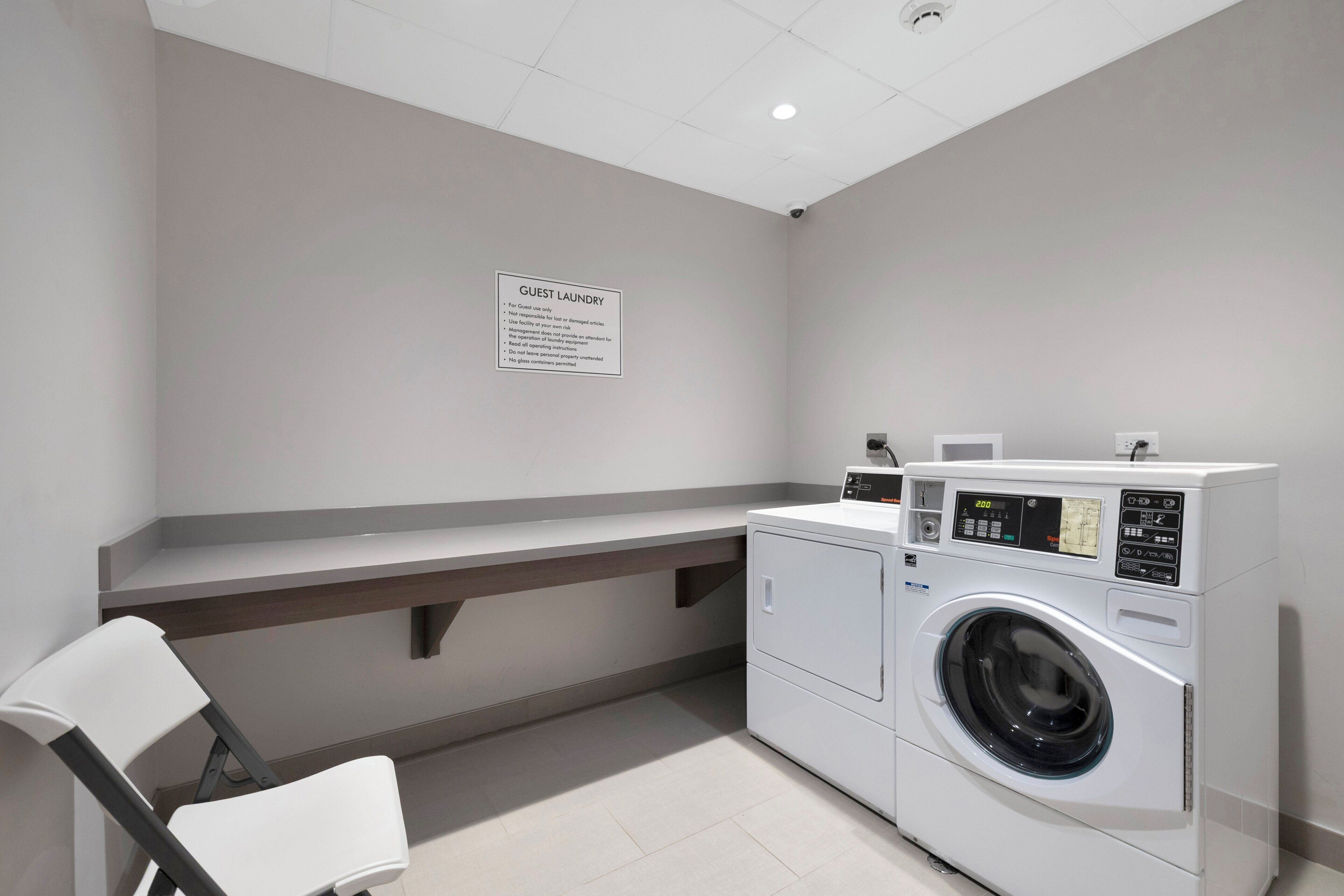 Een wasruimte met een bord dat aangeeft dat deze voor gasten is, een wasmachine, een droger en een stoel.