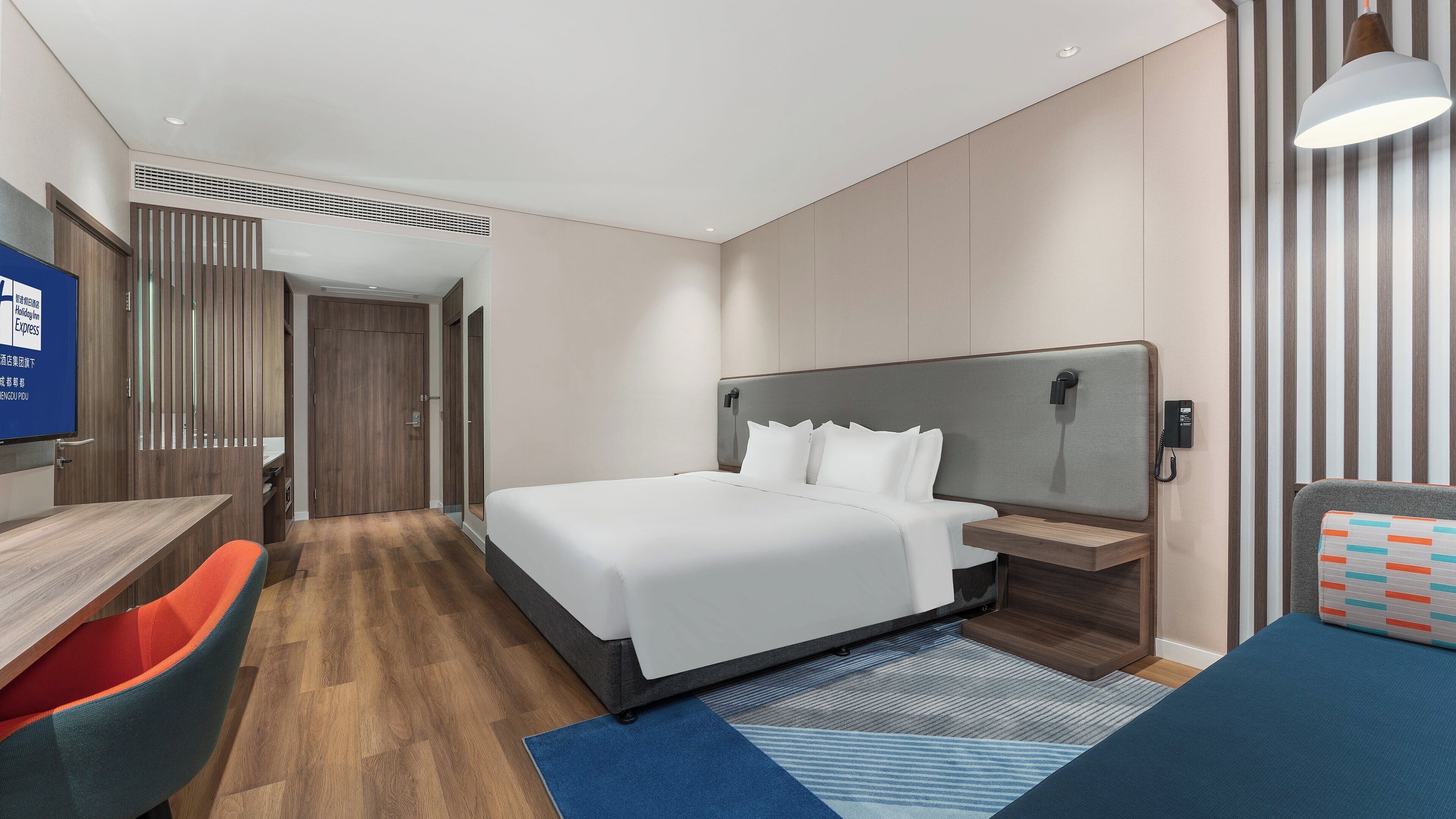 Ein modernes Hotelzimmer mit einem großen Bett, einem Schreibtisch und einem Sessel. Ein Fernseher ist an der Wand angebracht und ein Telefon steht auf dem Nachttisch.