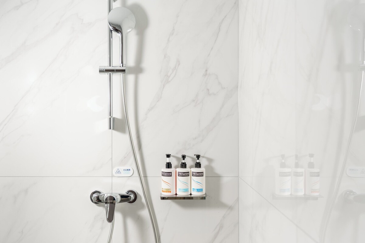 A bathroom with a shower, a hand dryer, and three bottles of shampoo.