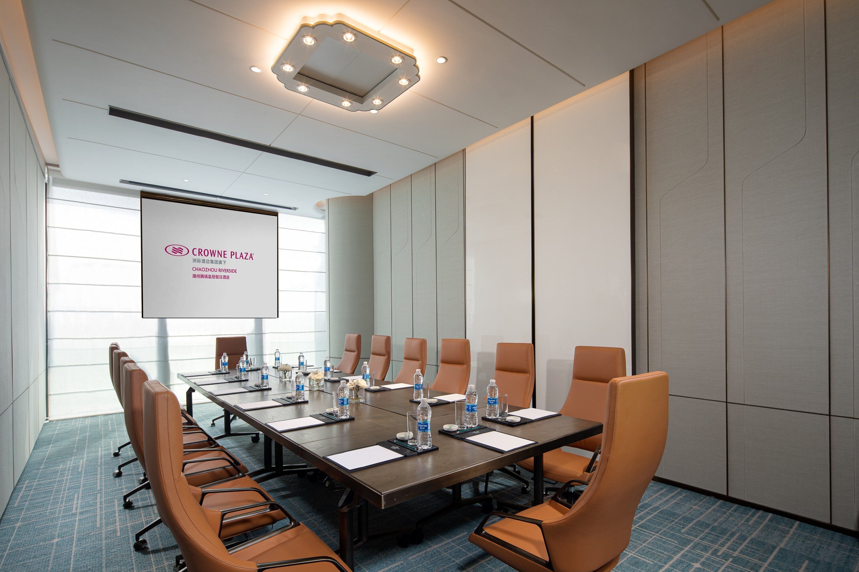 Sala de conferências com uma mesa grande, cadeiras, garrafas de água e uma tela de projeção.