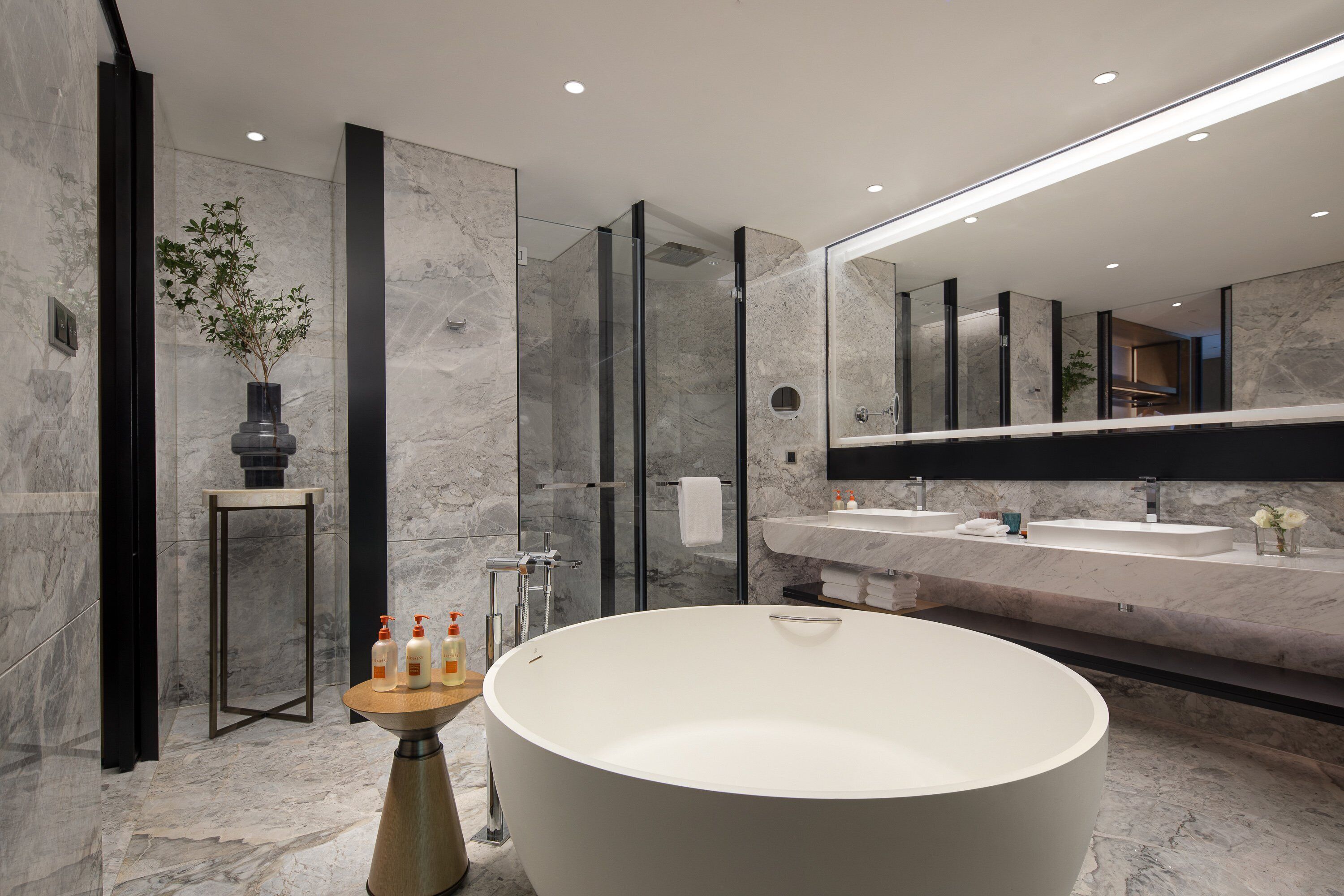 A modern bathroom with a freestanding bathtub, a large mirror, and a marble-tiled wall.