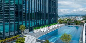 A modern building with a swimming pool, surrounded by greenery and white lounge chairs.