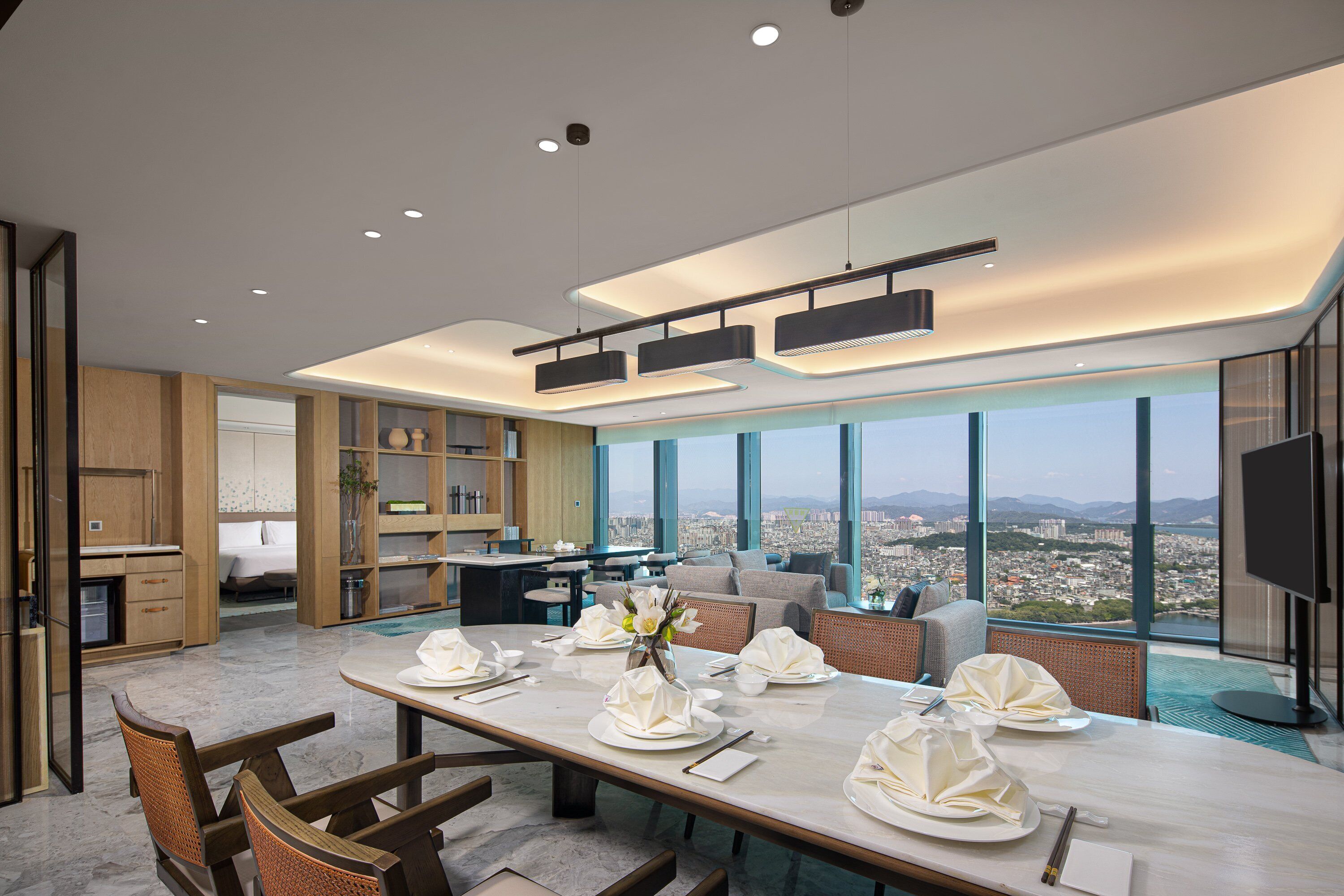 A modern dining area with a long table set for a meal, a kitchen in the background, and a city view through large windows.