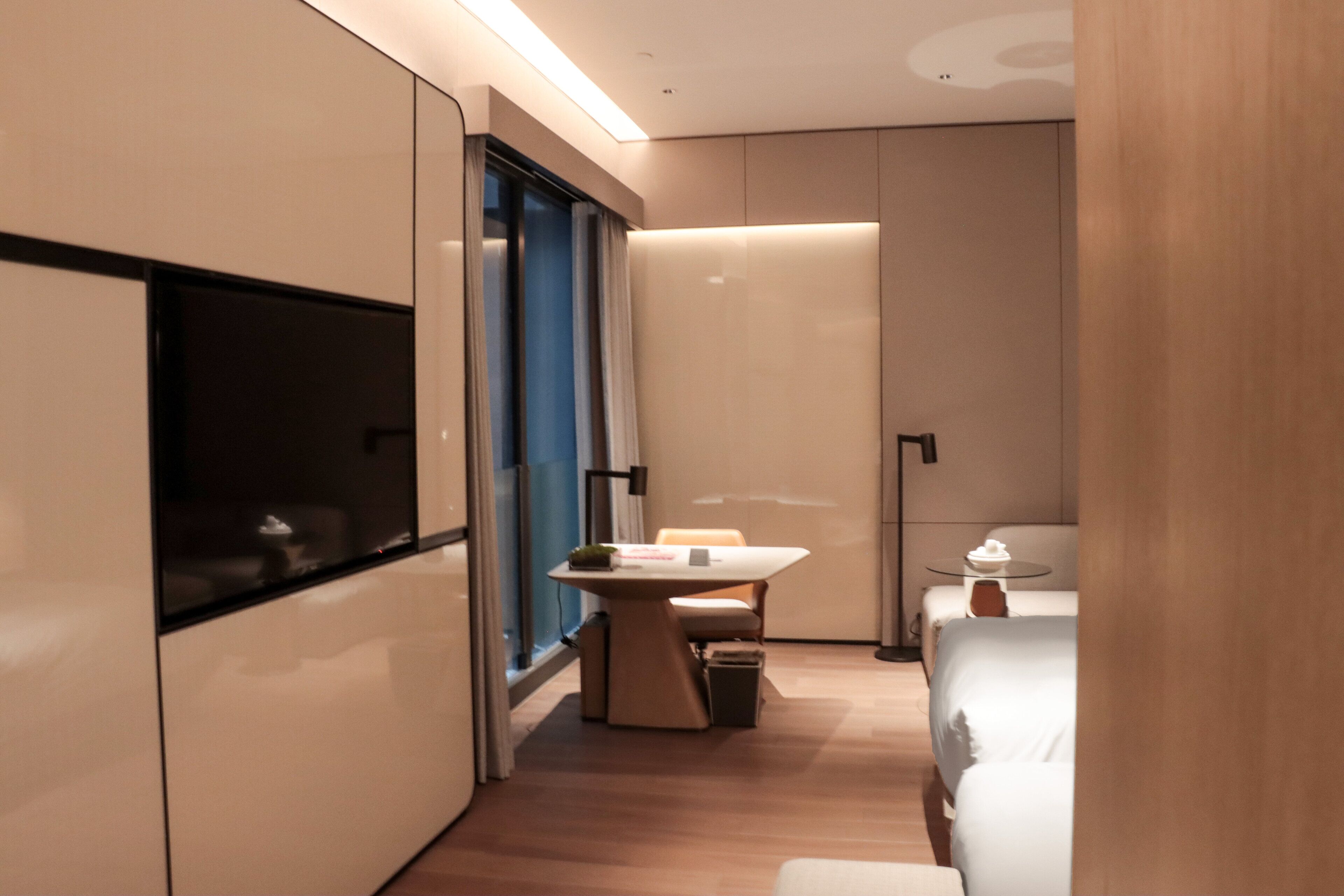 A modern hotel room with a bed, a desk, a chair, a small table, and a TV.