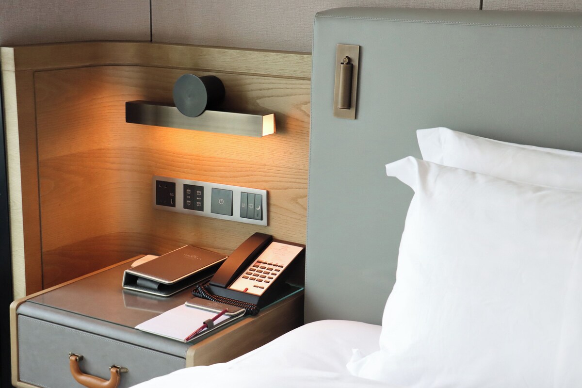 A hotel room with a wooden headboard, a bedside table with a phone and a notepad, and a suitcase.