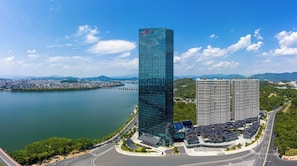 A modern high-rise building with a glass facade, adjacent to a body of water and a bridge, surrounded by greenery and other buildings. - Crowne Plaza Chaozhou Riverside by IHG (Chaozhou)