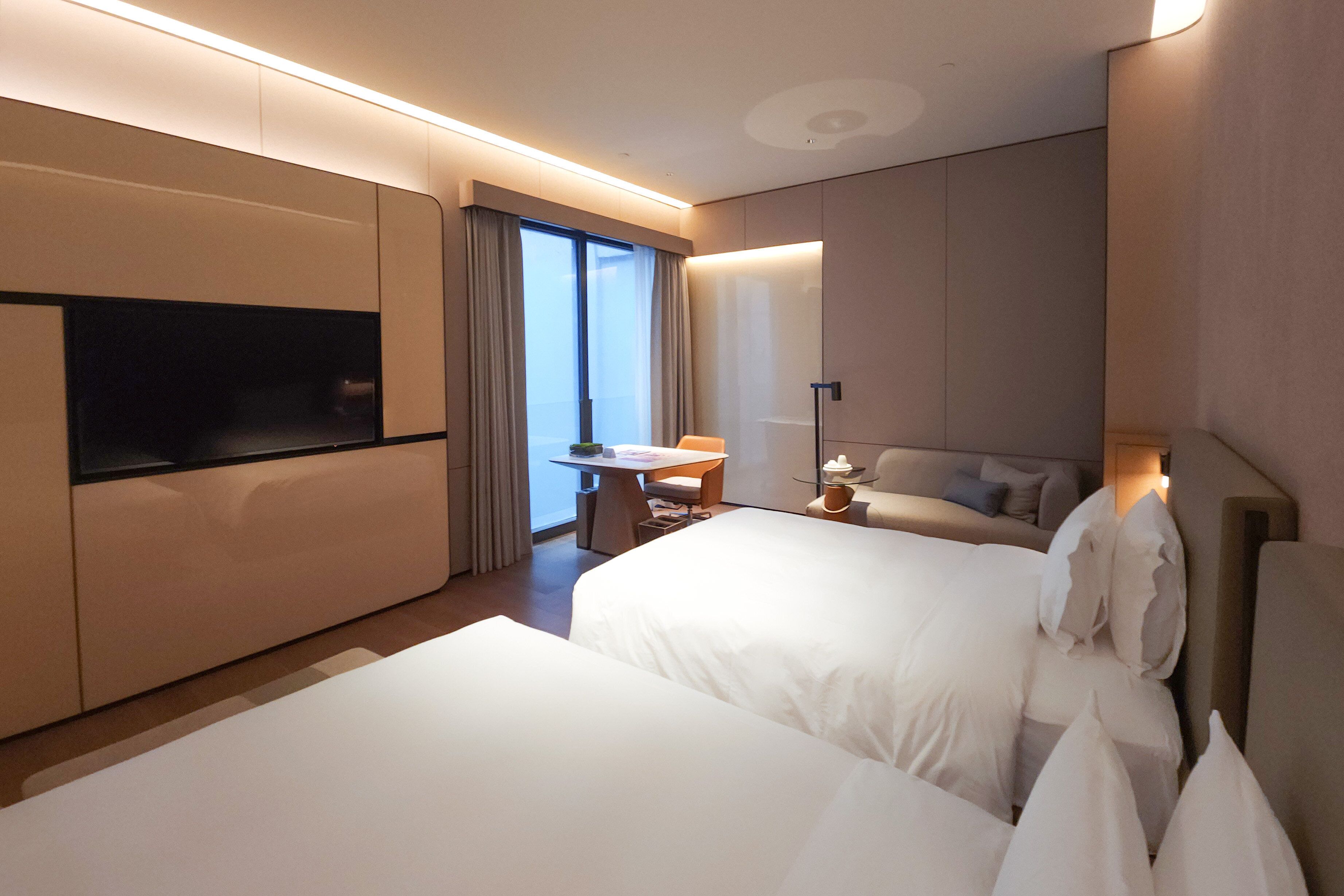 A hotel room with two beds, a TV, a small table, a lamp, and a view of the outdoors.