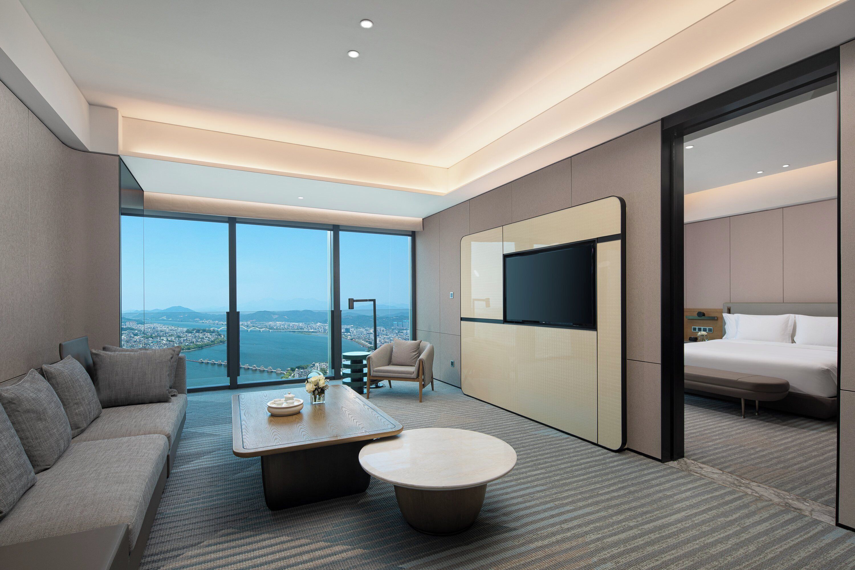 A modern hotel room with a large window offering a city view, a sofa, a coffee table, a TV, and a bed.