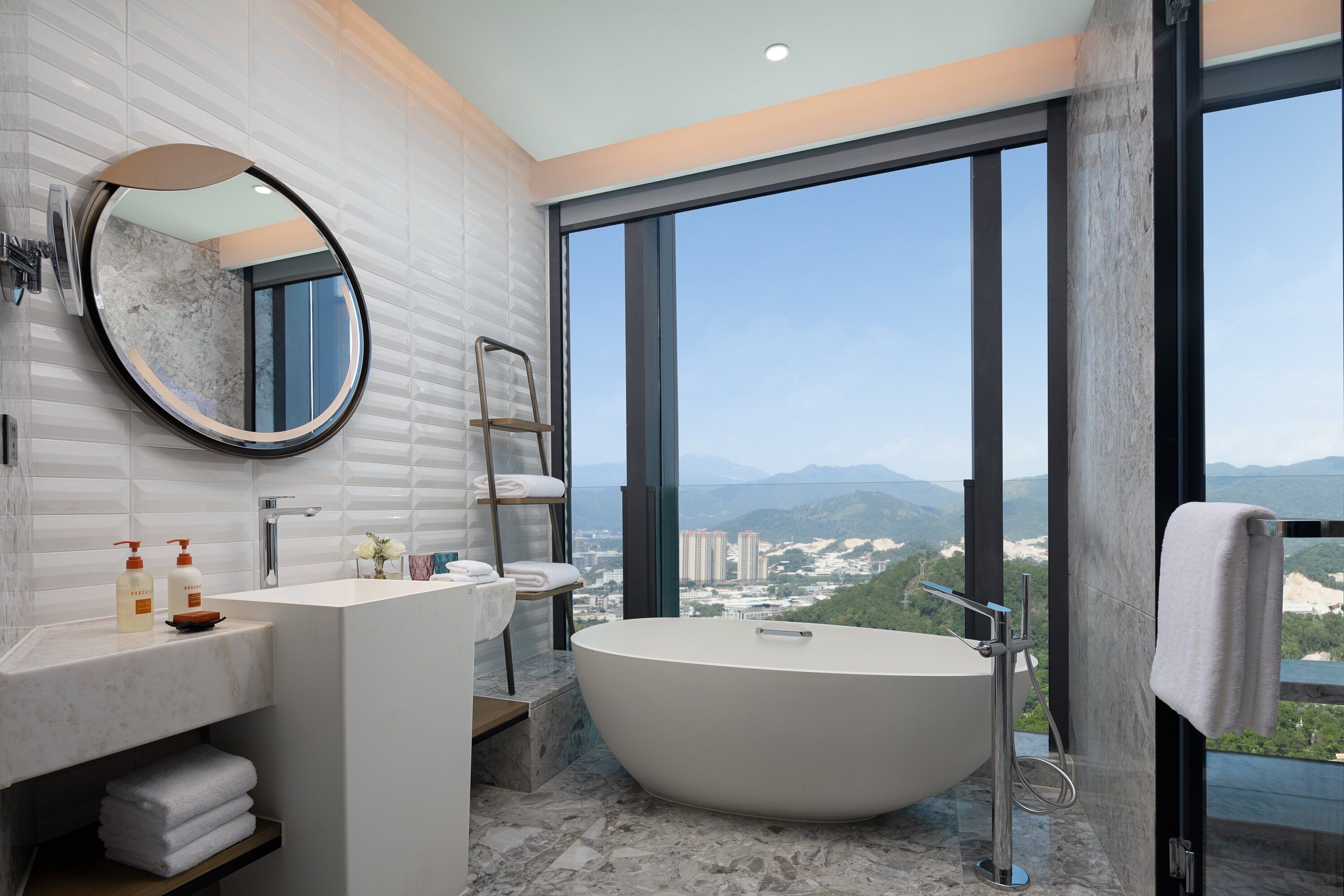 A modern bathroom with a freestanding bathtub, a large mirror, and a view of a cityscape through large windows.