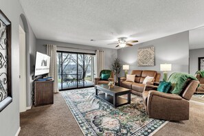 A living room with a sofa, coffee table, and a television. There is a large window with curtains and a view of the outdoors.