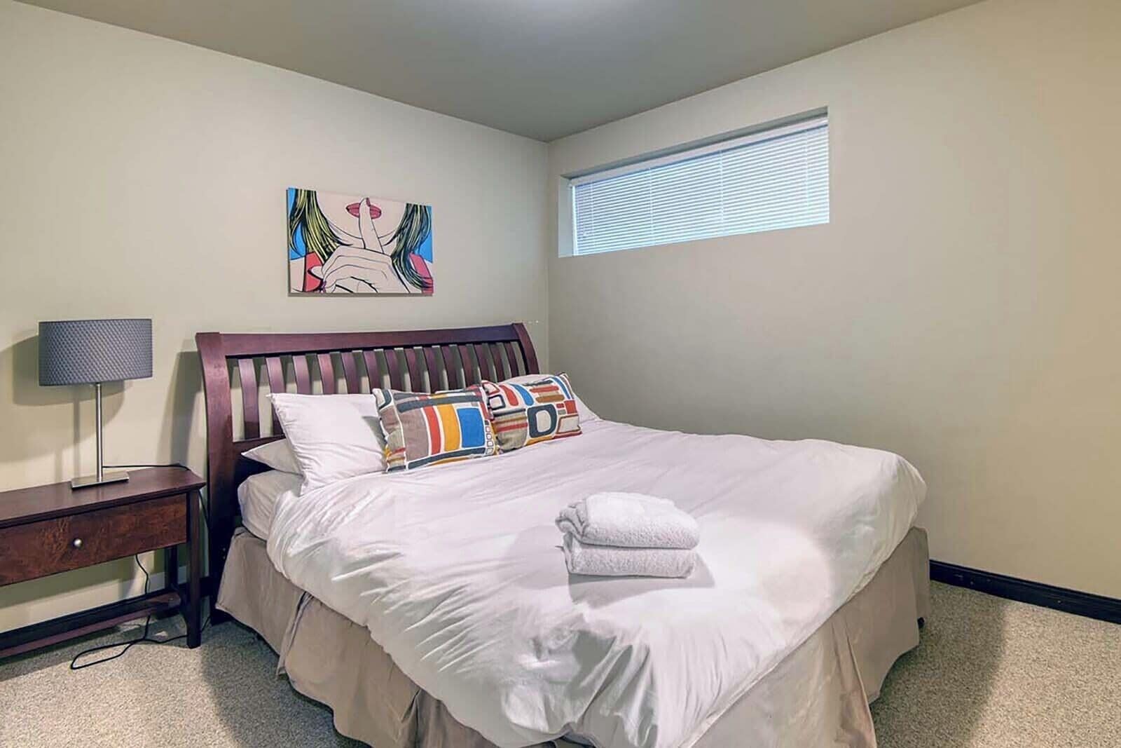 Un lit bien fait, avec une literie blanche et des coussins colorés, une table de chevet avec une lampe, une œuvre d’art accrochée au mur et une fenêtre avec des stores.