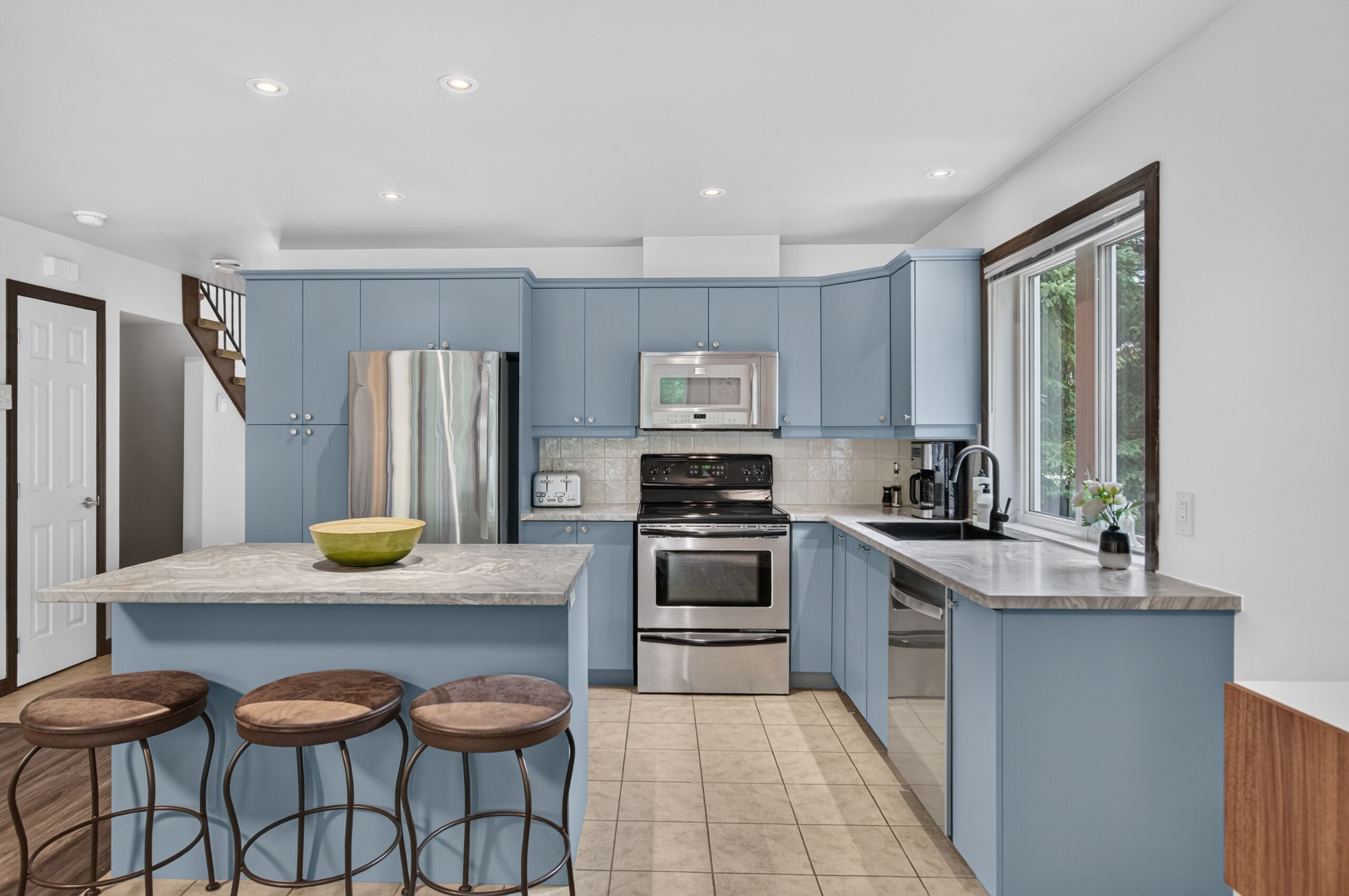 A modern kitchen with blue cabinets, a central island with stools, stainless steel appliances, and a window providing natural light.