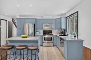 A modern kitchen with blue cabinets, a central island with stools, stainless steel appliances, and a window providing natural light. - Borealis Homes (Mont-Tremblant)