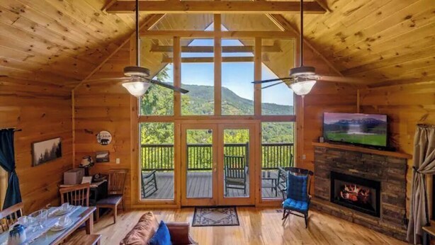Interior de una cabaña de madera con comedor, chimenea y un amplio ventanal con vistas a un paisaje montañoso.