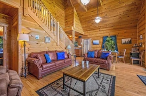 Un intérieur spacieux de chalet en bois, avec un escalier en bois, des canapés en cuir dotés de coussins bleus, une table basse en bois et un coin repas.