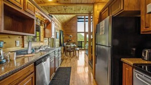 Interior de una cabaña de madera con cocina, comedor y vistas al exterior.