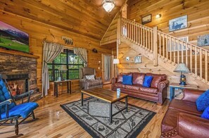 Interieur van een houten chalet met een open haard, leren banken, een houten trap en uitzicht op de natuur.