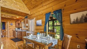 Interior de una cabaña de madera con comedor, cocina y un amplio ventanal con cortinas.