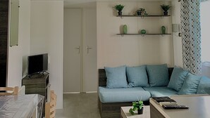 Uma sala de estar moderna com sofá, mesa de centro e televisão. Há prateleiras com plantas e uma porta que dá para outro cômodo.