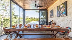 A dining area with a wooden table set for a meal, large windows, and artwork on the walls. - Villa LaGú • Luxury 5BR Jungle Villa • Pool, Spa & Chef (Tulum)