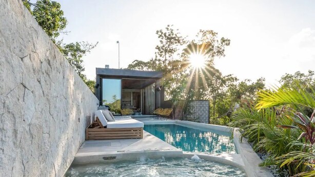 A modern outdoor pool area with a stone wall, lounge chairs, and a wooden deck. - Villa LaGú • Luxury 5BR Jungle Villa • Pool, Spa & Chef (Tulum)