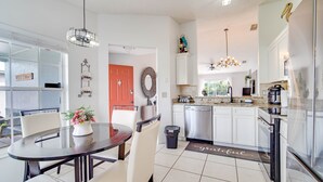 Una cocina moderna con armarios blancos, encimera de granito y electrodomésticos de acero inoxidable. Hay una zona de comedor con una mesa redonda y sillas, y una puerta roja que da a otra habitación.