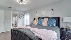 Een slaapkamer met een gebonden hoofdbord, een bed met een grijze deken en roze en blauwe kussens, een nachtkastje met een lamp, en een wand met ingelijste kunstwerken.