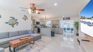 Un salon moderne avec un canapé, une table basse et un ventilateur de plafond. Il y a un coin cuisine équipé d’un four à micro-ondes et de placards. Une grande fenêtre offre une vue sur une plage avec des chaises longues.