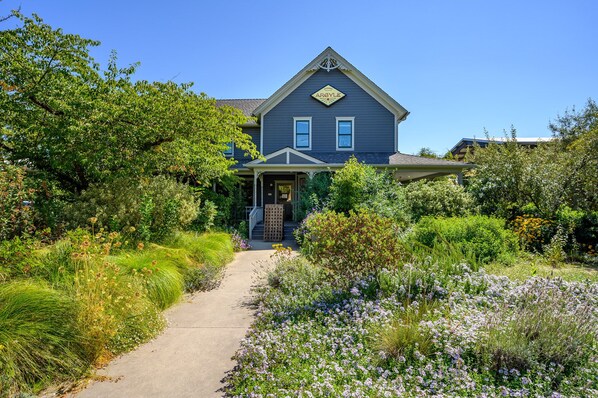 Une maison à deux étages, à l’extérieur d’un bleu foncé, avec un porche à l’avant et un jardin où poussent différentes plantes et fleurs.