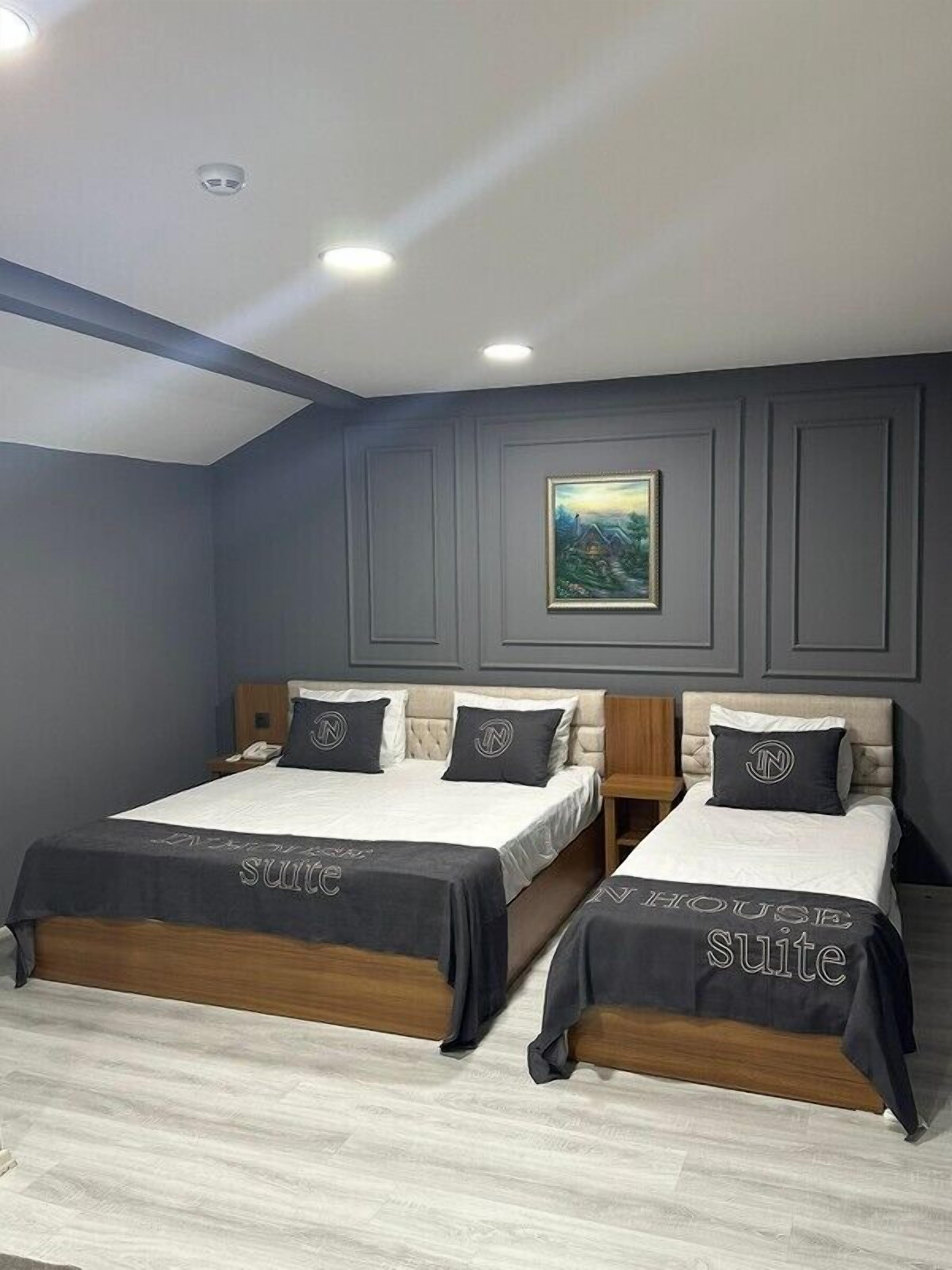 Quarto de hotel com duas camas, cada uma com cabeceira e mesa de cabeceira. O quarto possui uma luminária no teto e um quadro na parede.
