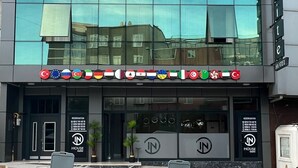 Un hôtel moderne, avec une façade en verre réfléchissant, une grande enseigne indiquant le nom « HOUSE » et une série de drapeaux signalant sa présence à l’échelle internationale.