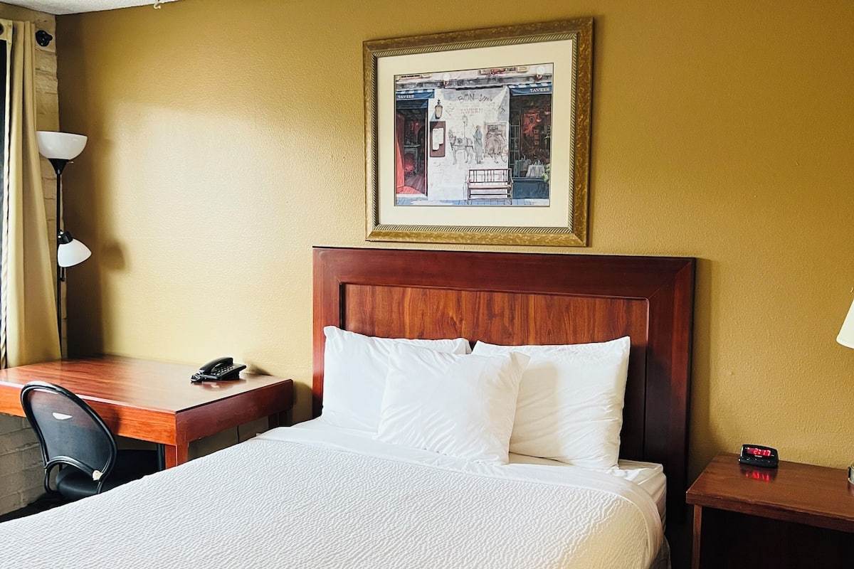 A hotel room with a bed, a desk with a telephone, a nightstand with a clock, and a framed picture on the wall.