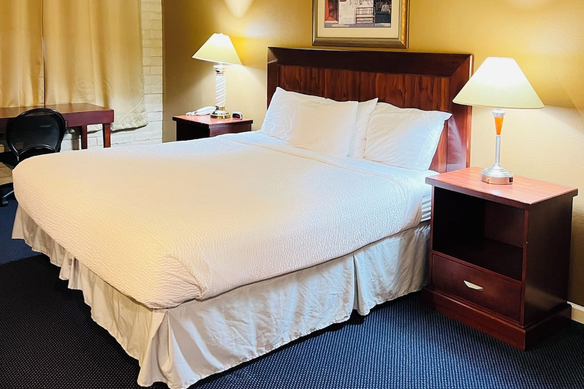 A hotel room with a large bed, two bedside tables with lamps, a wooden headboard, a framed picture on the wall, and a chair.
