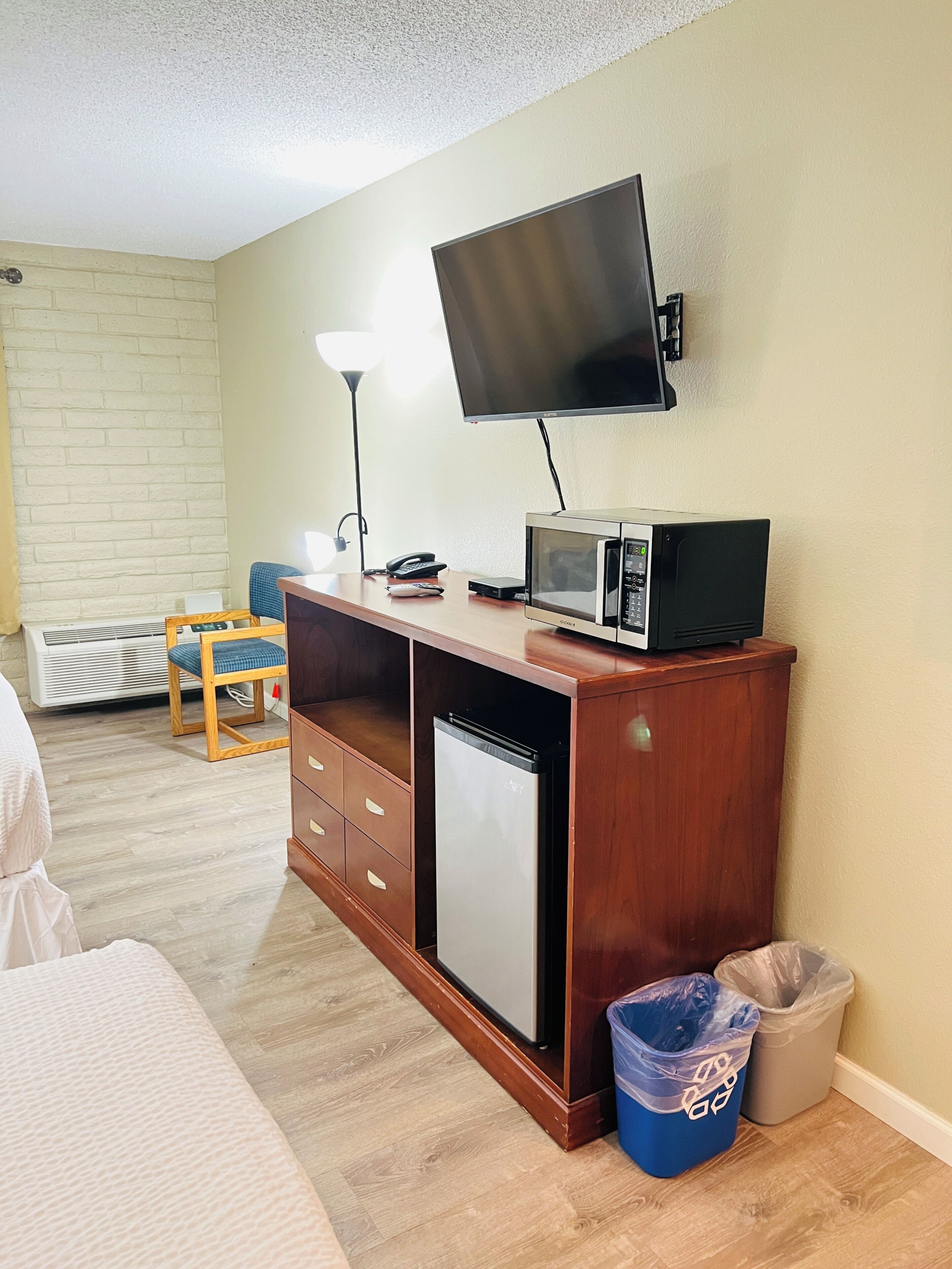 Habitación compacta con escritorio de madera, televisión de pantalla plana, microondas, mini refrigerador, silla, lámpara y un basurero.