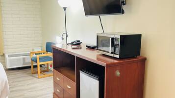 Habitación compacta con escritorio de madera, televisión de pantalla plana, microondas, mini refrigerador, silla, lámpara y un basurero.