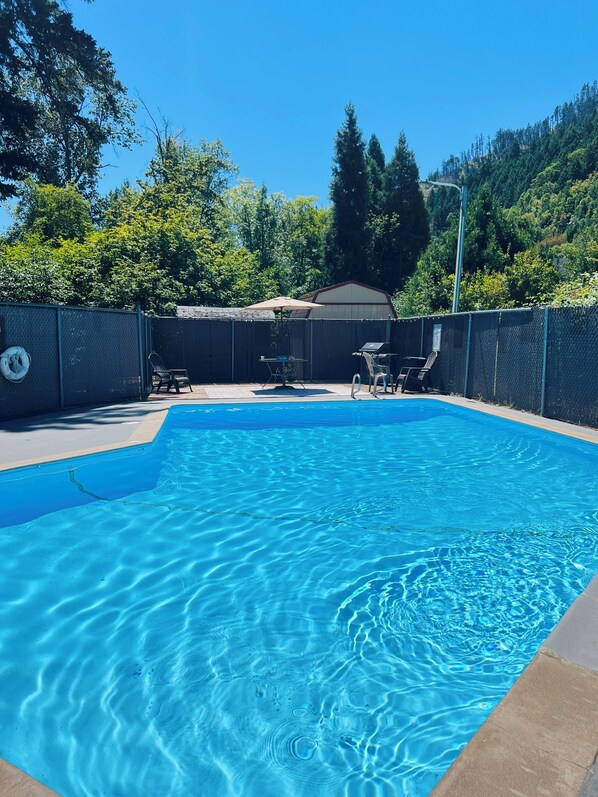 A clear blue swimming pool with a fenced area, a covered seating area, and trees in the background.