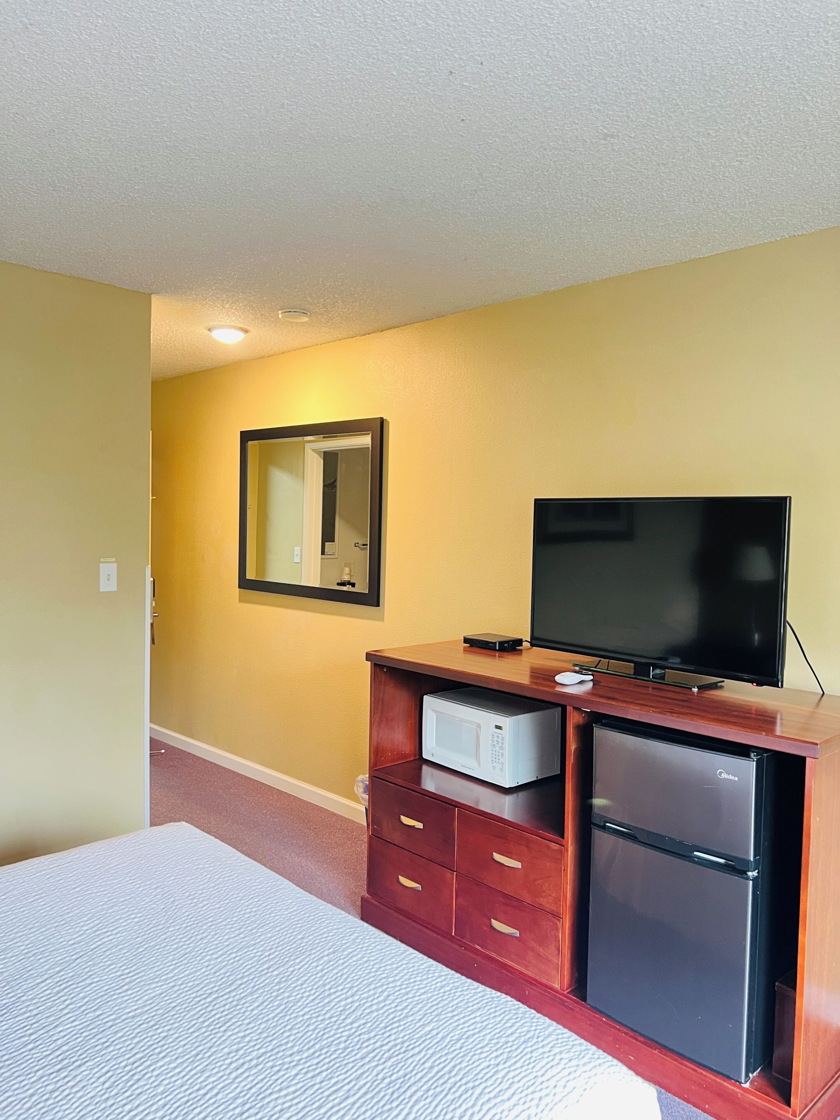 A hotel room with a bed, a wooden dresser with a television and a microwave, a mirror, and a door leading to another room.