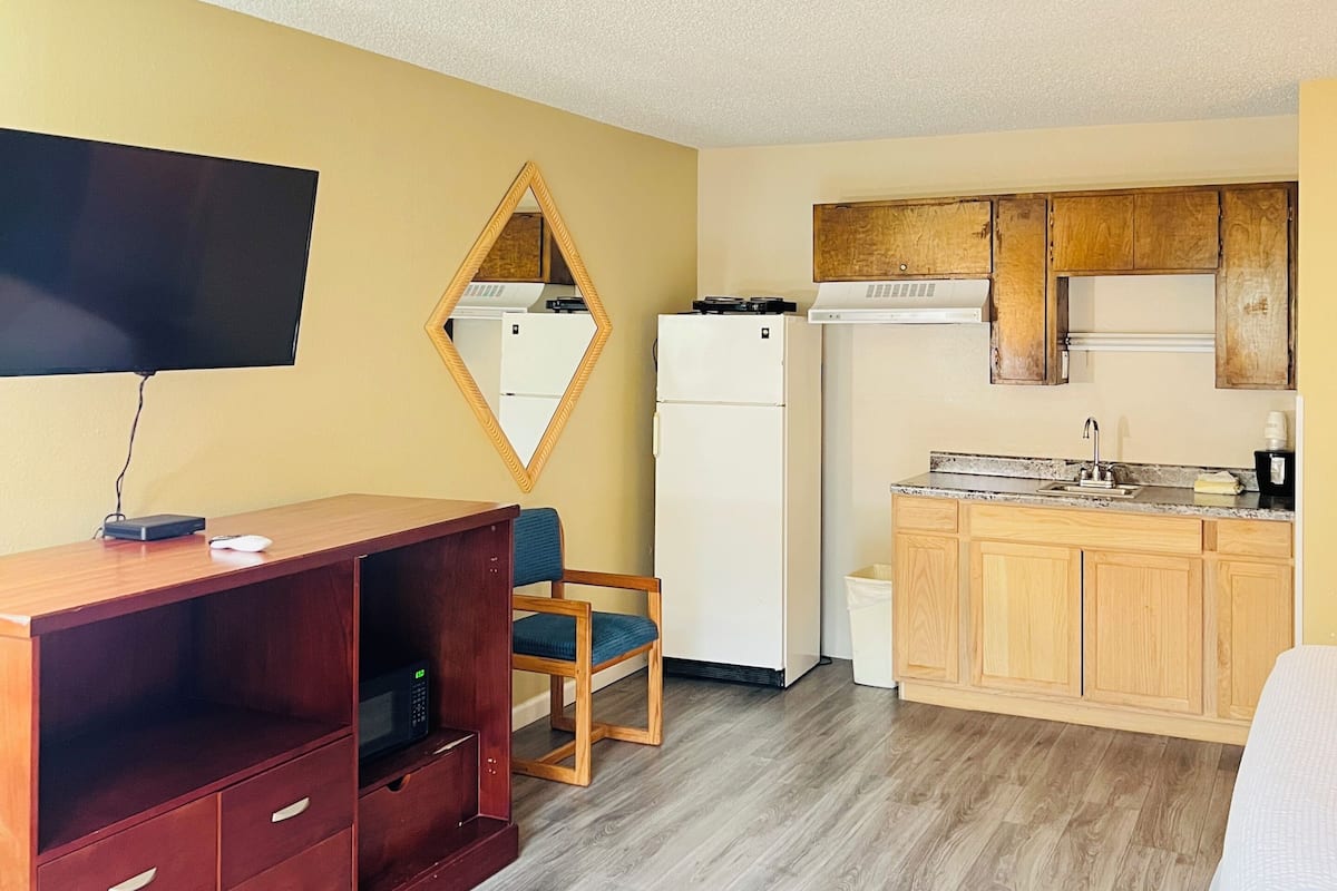 A compact room with a wooden desk, a chair, a flat-screen TV, a refrigerator, a microwave, and a sink.