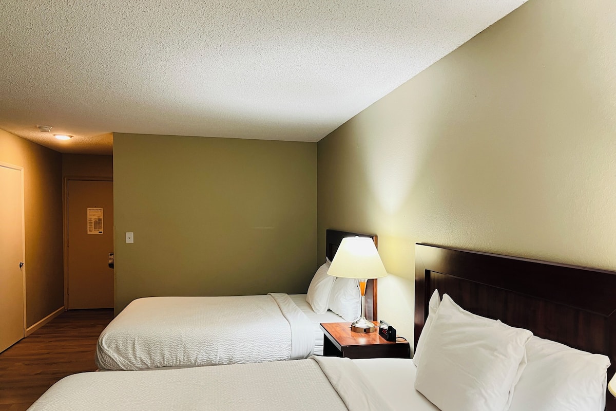 A hotel room with two beds, a nightstand with a lamp, a wooden headboard, and a door leading to another room.