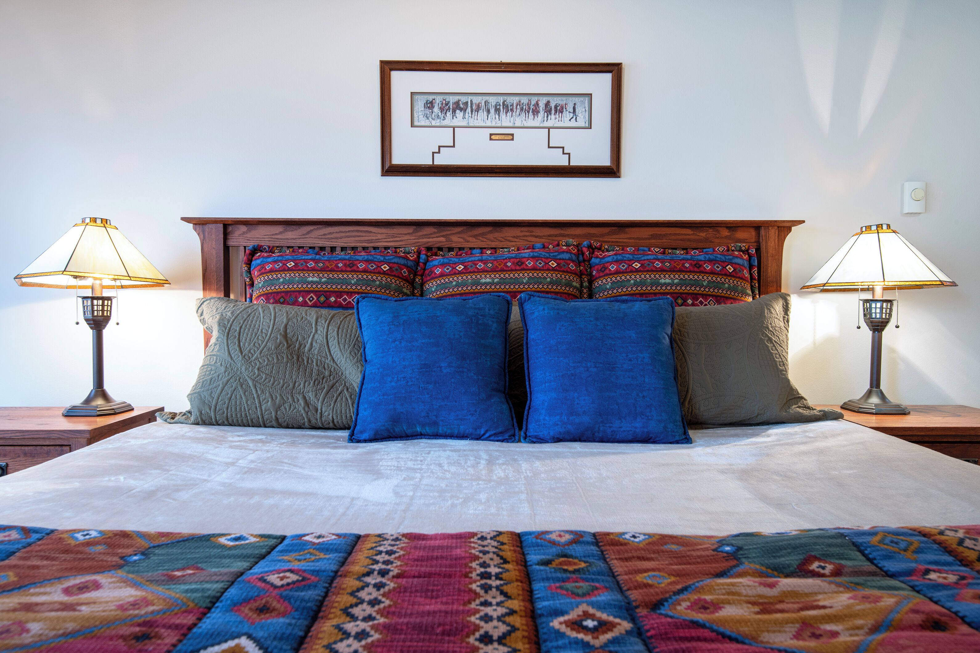 A neatly made bed with a patterned bedspread, a wooden headboard, and multiple pillows. There are two bedside lamps on either side of the bed.