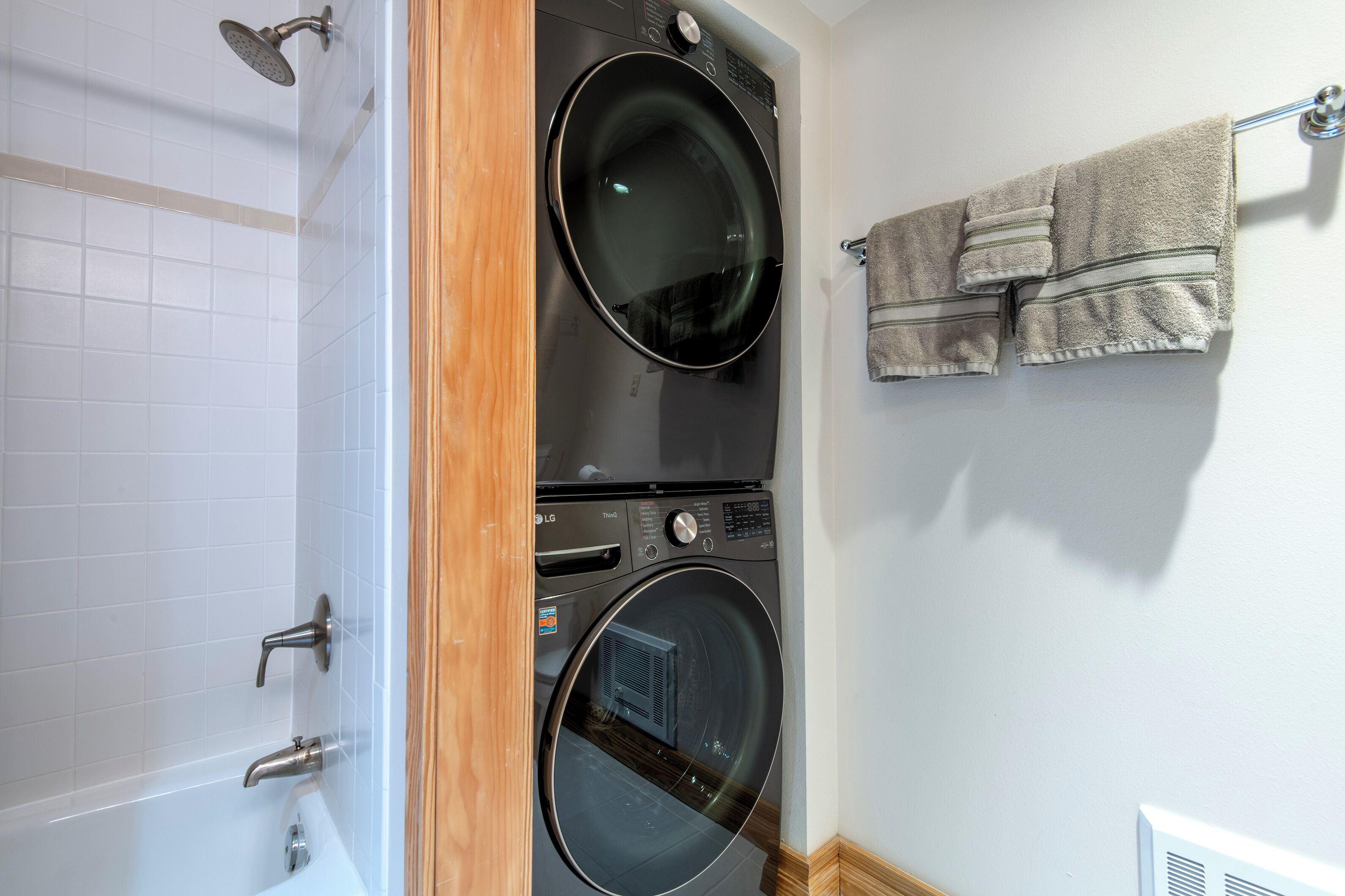 A laundry area with two stacked front-loading washing machines, a white tiled shower area, and a towel rack with towels.