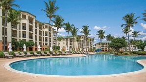 A resort pool with lounge chairs and umbrellas, surrounded by palm trees and hotel buildings.