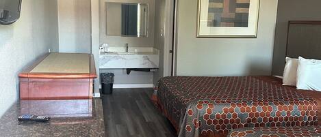 Una habitación de hotel con piso de madera, una cama con cubrecama estampado, una mesita de noche, un espejo, un cuadro enmarcado en la pared y un televisor montado en la pared.