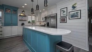 A modern kitchen with a central island, white cabinetry, teal accents, stainless steel appliances, pendant lighting, and framed artwork on the walls.