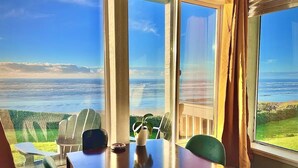 Une pièce dotée d’une grande fenêtre donnant sur un paysage magnifique, avec vue sur l’océan et les collines. On y trouve une table avec des chaises et une plante en pot.
