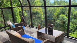 Une pièce meublée de mobilier en osier, avec une petite table et une vue sur une végétation luxuriante, visible à travers de grandes fenêtres.