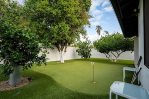 A well-maintained artificial grass golf course with a white wall and trees, a number 2 marker, and a white table with chairs.